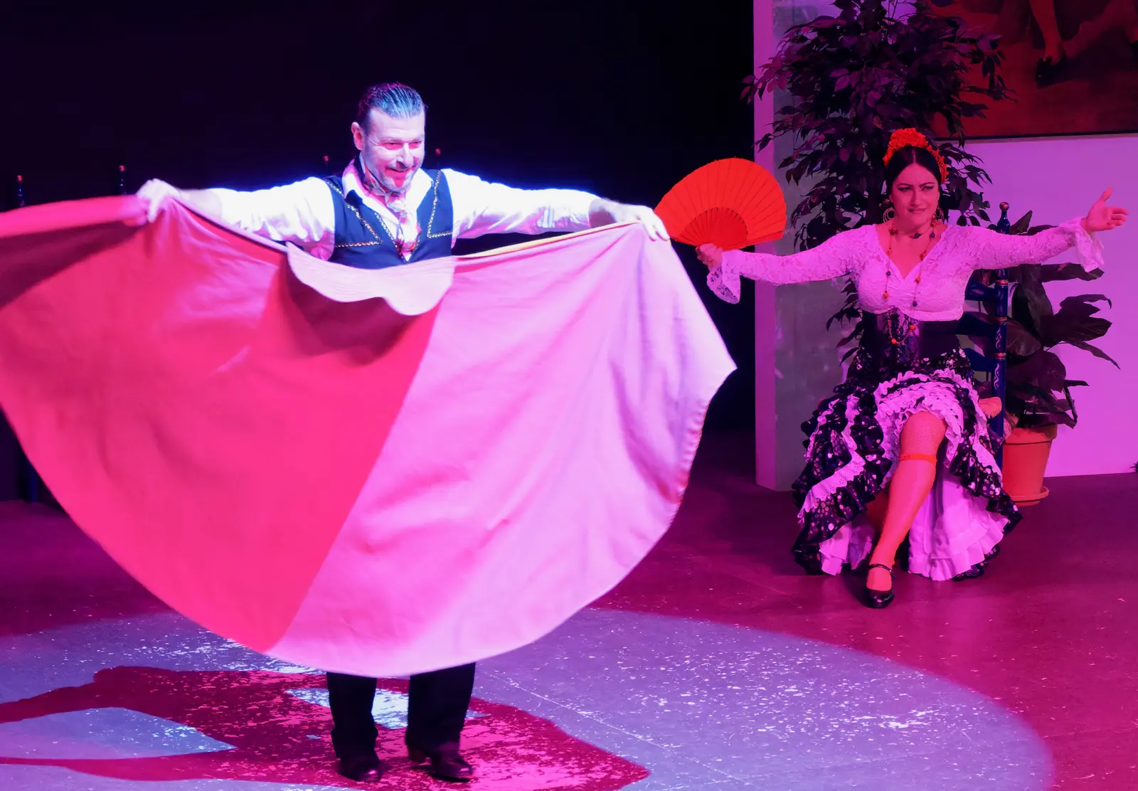 A male dancer holds out a wide pink and orange cape beside a seated dancer with a red fan