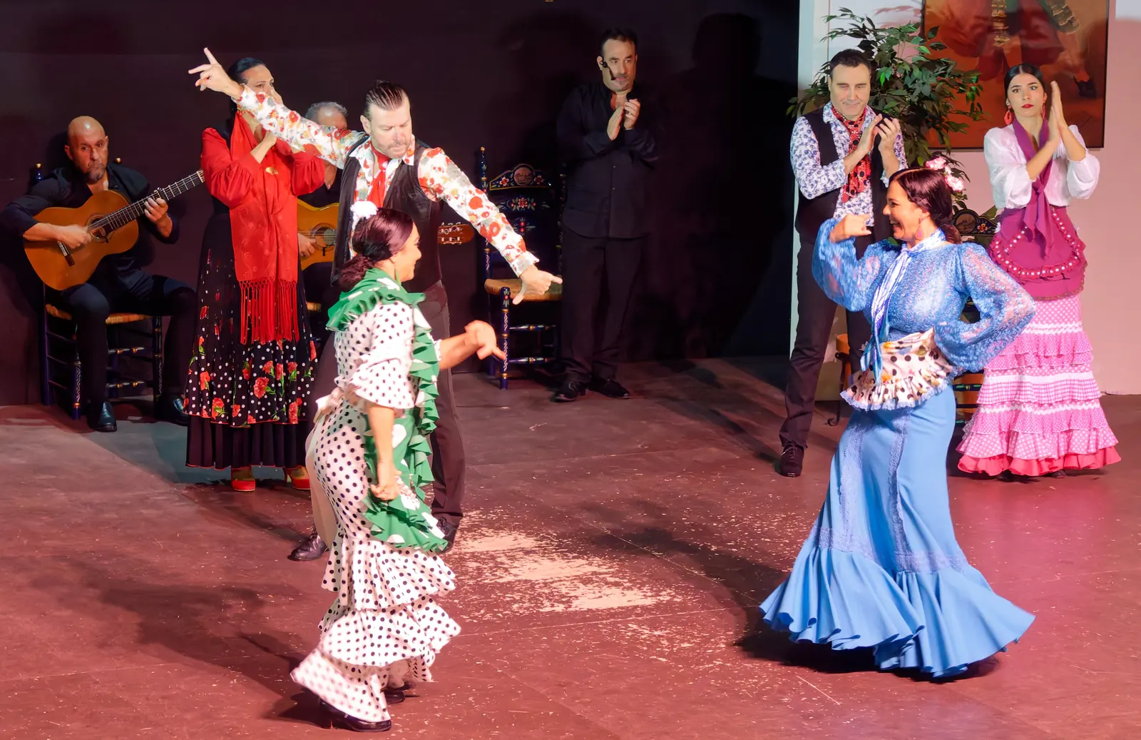 Dancers in blue, pink, green and red dresses move across the stage while guitarists play behind them