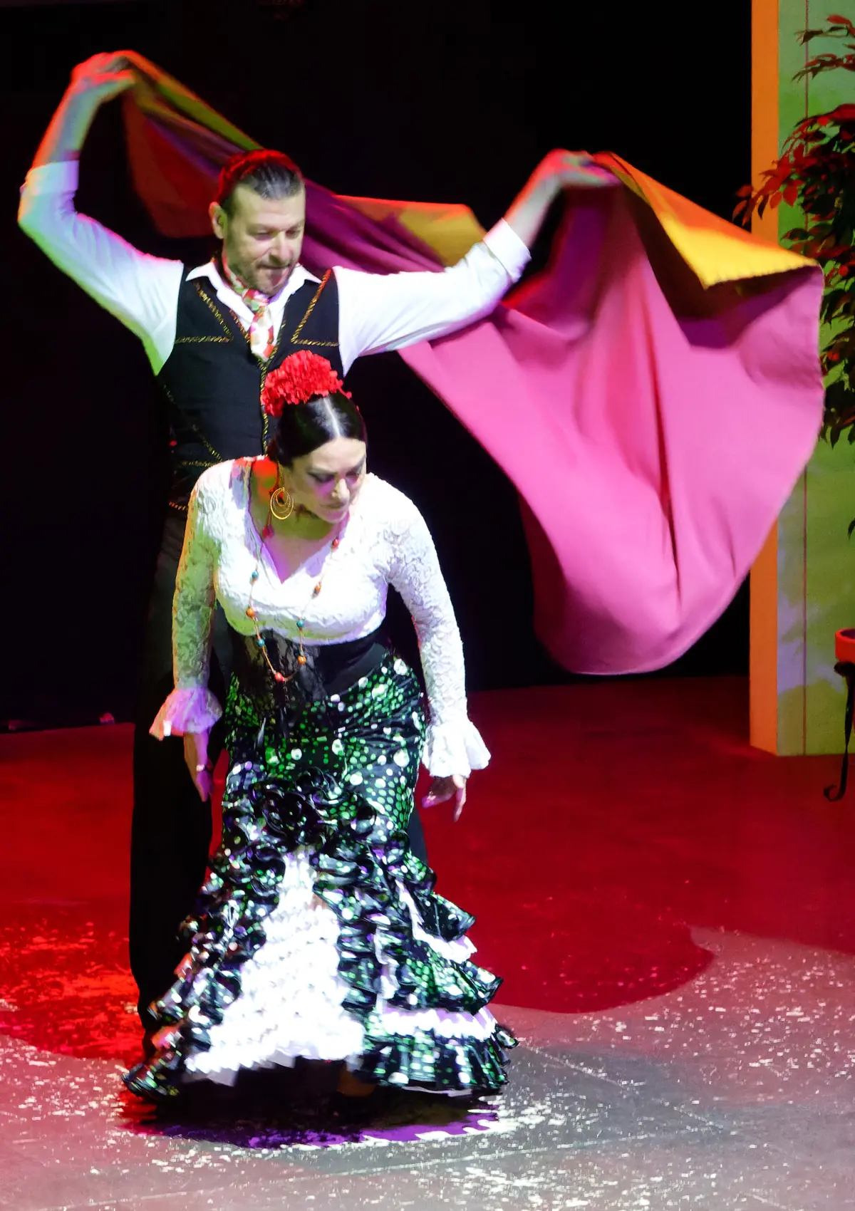 A dancer in a black polka-dot dress and a dancer in a white blouse and ruffled skirt perform together on a red-lit stage
