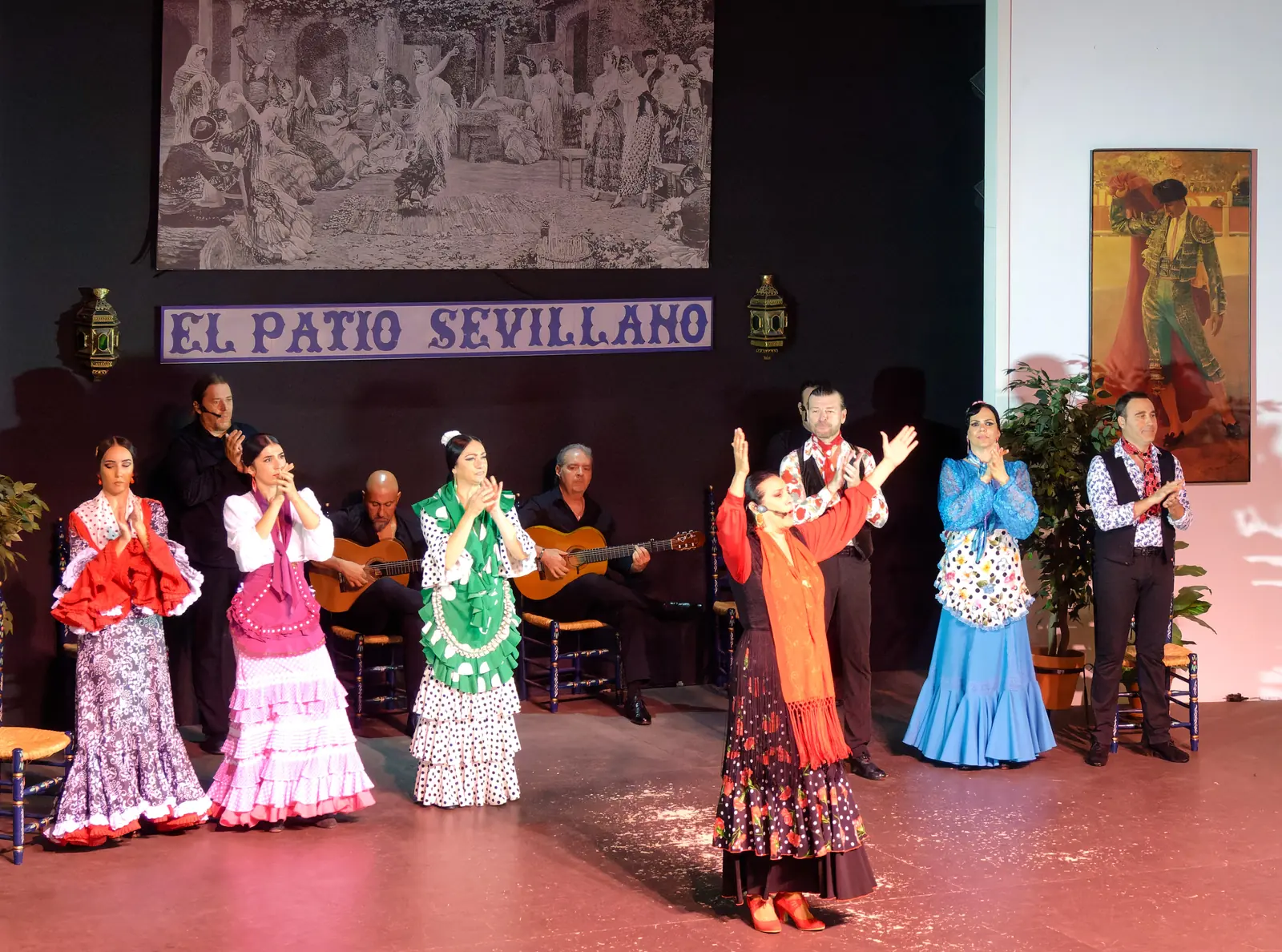 The whole company standing and clapping on the stage, dancers in polka-dot and ruffled dresses with two guitarists seated behind, beneath a blue tile sign reading El Patio Sevillano
