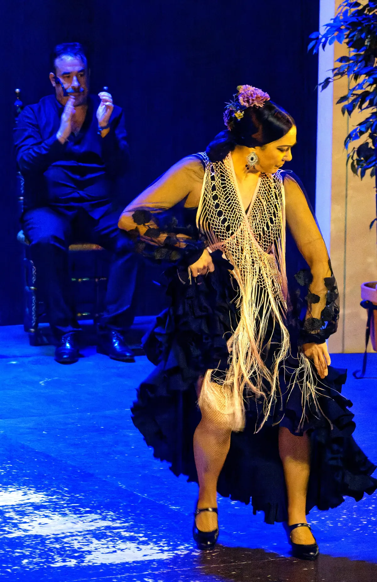 A dancer in a black ruffled dress and a long cream fringed shawl on a blue-lit stage, with a seated singer behind