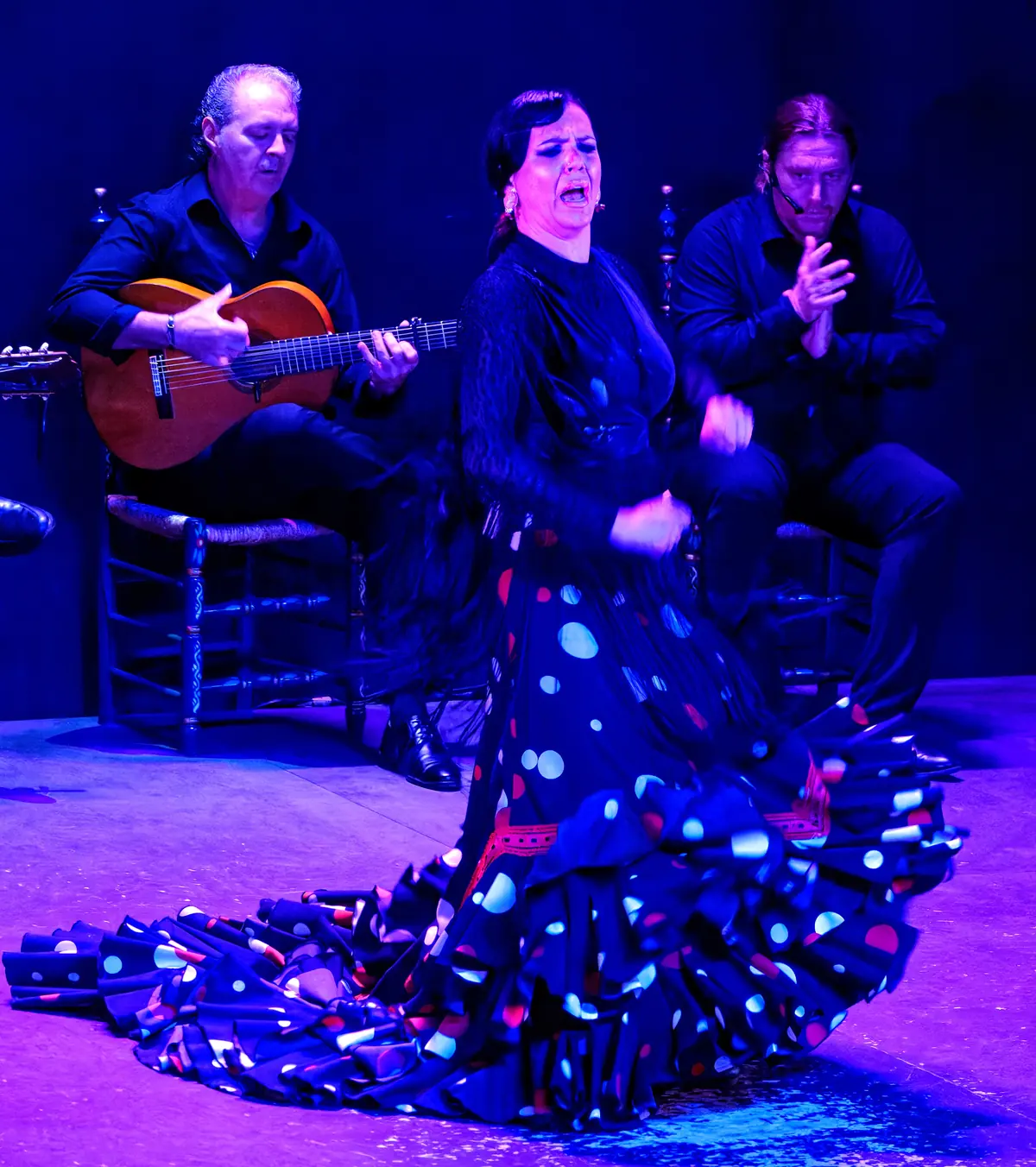A dancer in a blue dress with white polka dots in front of a guitarist and a clapping artist, under purple light