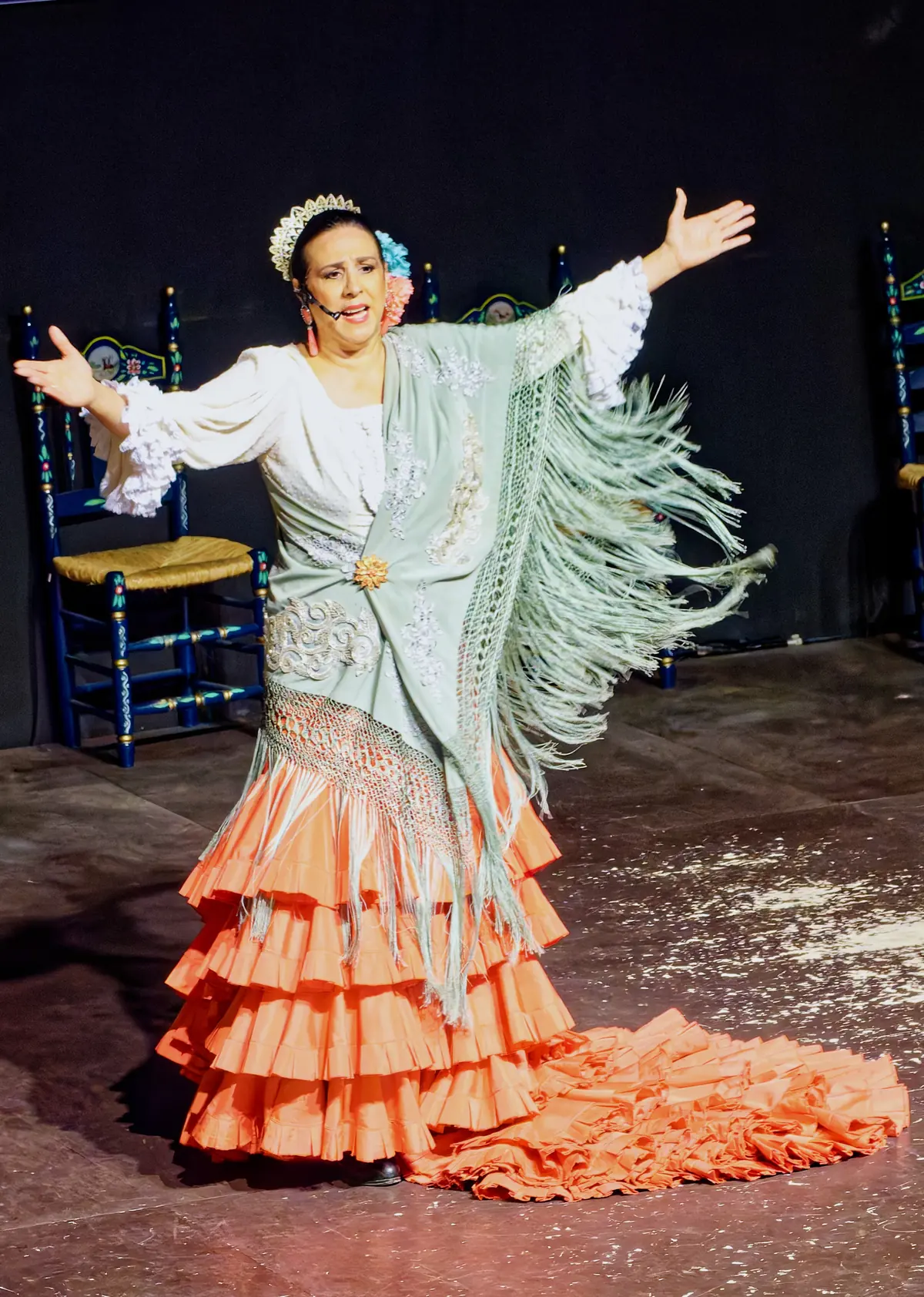A dancer in a peach ruffled dress and a pale green fringed shawl with her arms spread wide on a dark stage